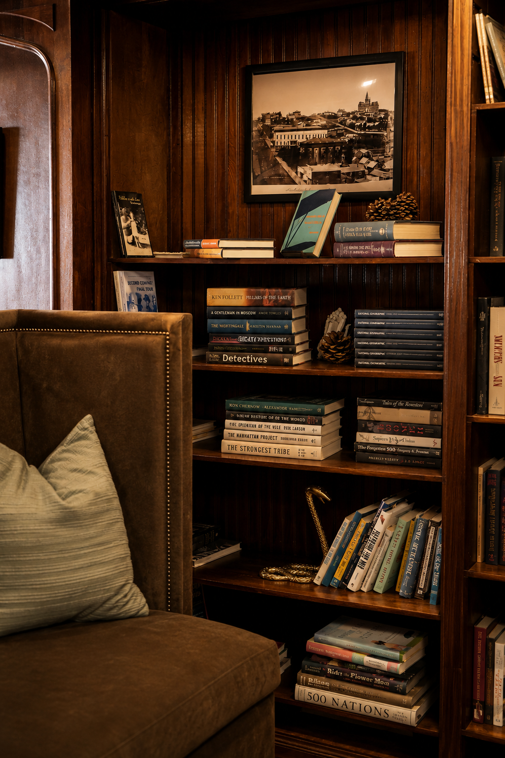 Stacked books and a framed historic Omaha photograph on the mahogany shelves beside an upholstered chair at The Reading Room